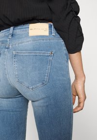 Modré denimové džíny s přiléhavým střihem, dvěma zadními kapsami a koženou nášivkou s logem na pásku. Hladká textura, střední odstín. 