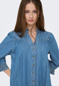 Νεαρή γυναίκα με μακριά καστανά μαλλιά, φορεμένη με ένα κουμπωτό πουκάμισο από τζιν με μανίκια τριών τετάρτων, στέκεται με τα χέρια στη μέση μπροστά σε ένα απλό φόντο.