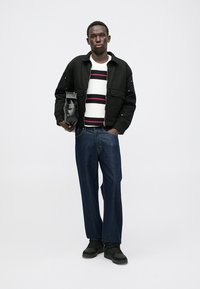 Juoda striukė su kišenėmis, balta ir tamsiai mėlyna juostuota megztinis, mėlynos džinsus ir juodus sportbačius; laiko mažą juodą maišelį.