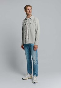 Grijze button-up overhemd met twee zakken over een wit en grijs gestreept T-shirt, gecombineerd met lichtblauwe jeans en witte sportieve schoenen.