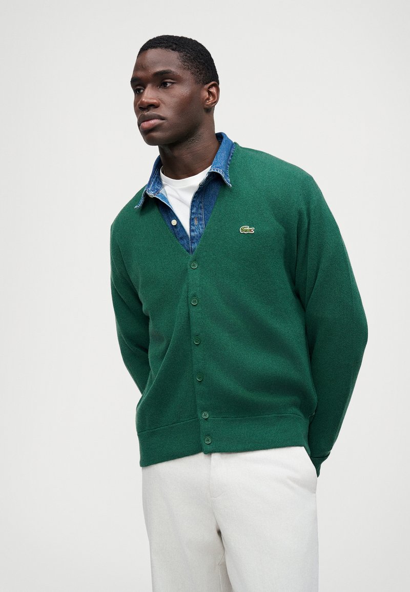 Cárdigan de punto verde con escote en V, cierre de botones y logo bordado, superpuesto a una camisa de denim. Pantalones blancos completan el look.