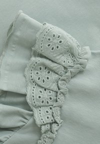 Lys grønn stoff med teksturerte volanger i broderte blomsterdesign og små utskjæringer langs kanten. Myk drapering og glatt overflate.