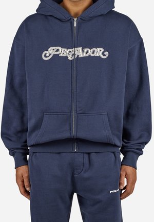Sudadera con capucha azul marino con cremallera y texto "PEGADOR" en el pecho, bolsillo canguro, puños acanalados, combinada con pantalones deportivos azul marino a juego que llevan un pequeño logo.