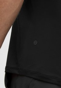 Črna športna majica z teksturirano površino, z zaobljenim robom in majhnim črnim logotipom na strani. Poglej bližje na tkanino in zasnovo.