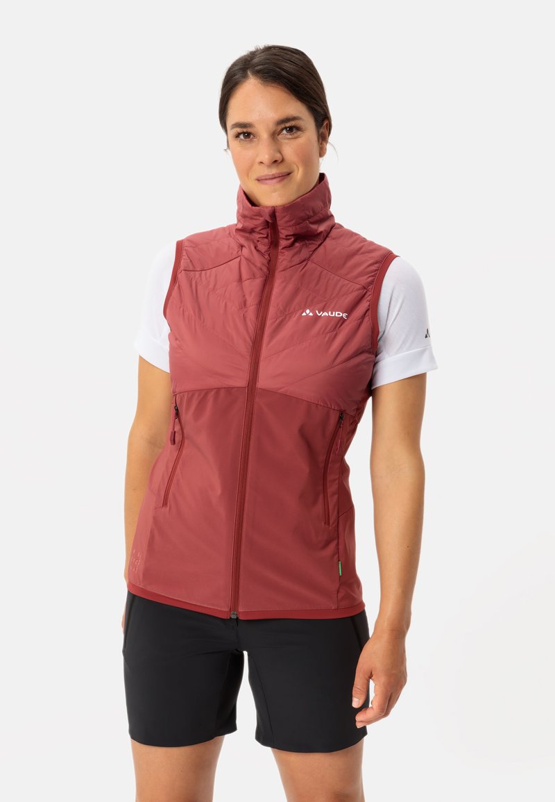 Gilet isolante rosso con collo alto, zip frontale e tasche laterali. Indossato sopra una camicia bianca a maniche corte, abbinato a pantaloni corti neri.