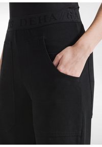 Zwarte broek met een brede tailleband voorzien van reliëftekst. Zijzakken met rechthoekige openingen en een gladde, matte textuur.