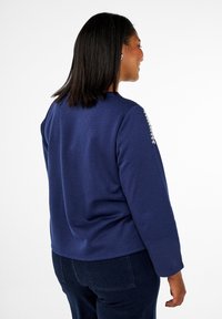Mörkblå långärmad topp med rund halsringning och dekorativa sömnadsdetaljer på axeln, tillverkad av ett mjukt, strukturerat tyg.