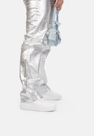 Pantalones metálicos plateados con un acabado brillante, combinados con zapatillas blancas y un pequeño bolso azul claro. Costuras detalladas en los pantalones.
