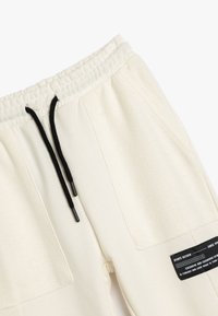 Cremefarvede sweatpants med elastisk talje og sorte snørebånd, struktureret stof og et fremtrædende sort mærke på siden.