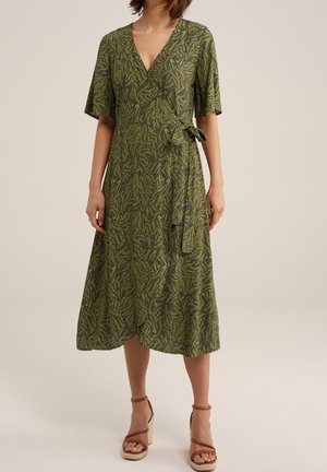 Vihreäkuvioinen kietaisumekko, jossa on lyhyet hihat ja vyötärönauhakiinnitys, puettuna ruskeiden korkeakorkoisten sandaalien kanssa neutraalilla taustalla.