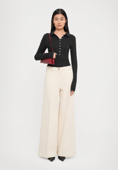 Femeie îmbrăcată cu un top negru cu mânecă lungă și nasturi, pantaloni largi de culoare crem, pantofi negri cu vârful ascuțit și purtând o geantă roșie de umăr.