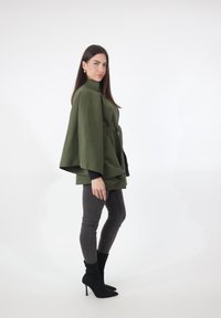 Olivgroene manteljas in cape-stijl met hoge kraag, ceintuurlijn en brede mouwen, gecombineerd met donkere nauwsluitende jeans en zwarte enkellaarsjes.