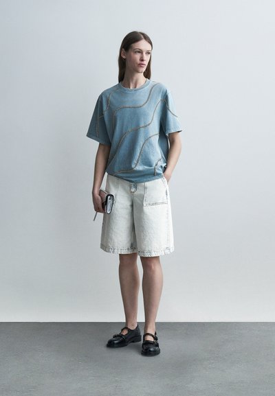 Νεαρή γυναίκα που φοράει ένα μπλε, φαρδύ t-shirt με ραμμένα σχέδια, λευκά σορτς μέχρι το γόνατο, μαύρα παπούτσια, κρατώντας μια μικρή μαύρη τσαντούλα.