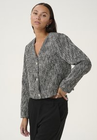 Kobieta o długich ciemnych włosach, ubrana w czarno-biały teksturowany sweter na guziki i czarne spodnie, pozująca z ręką na biodrze na tle jednolitego tła.