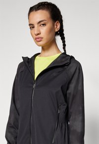 Schwarze leichte Zip-Jacke mit Mesh-Ärmeln, verstellbarem Bund und auffälligem Logo. Über einem leuchtend gelben Shirt getragen.