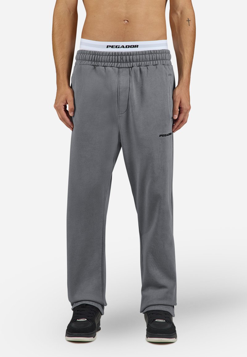 Pantaloni de trening gri cu o croială relaxată, talie elastică cu logo-ul "PEGADOR" și buzunare laterale. Purtați cu pantofi sport negri.