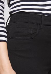 Jeans pretos elásticos com uma textura suave. Apresenta um pequeno bolso traseiro e uma cintura limpa, combinados com uma blusa de manga longa riscada.