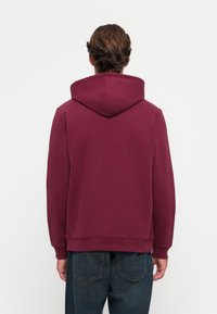 Bordeaux hættetrøje med snoretræk i hætten, ribbede manchetter og kant; blødt stof; ensfarvet design; båret med mørke jeans.