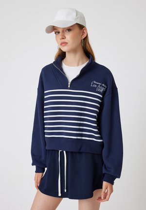 Sudadera corta de color navy con rayas blancas, cuello con cremallera y detalle bordado "Los Ángeles 1998". Combinada con una falda navy a juego.