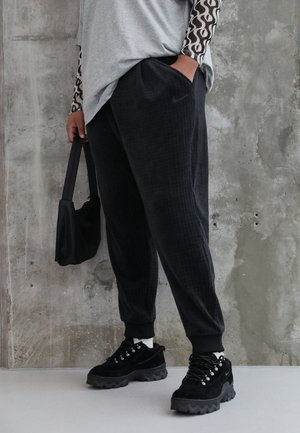 Person wearing black Nike joggers, black chunky sneakers, gray top with patterned sleeves, holding small black handbag against concrete wall.