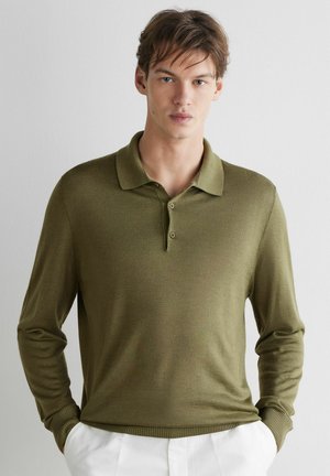 Giovane uomo con capelli castani corti che indossa una polo verde oliva a maniche lunghe e pantaloni bianchi, in piedi davanti a uno sfondo semplice.