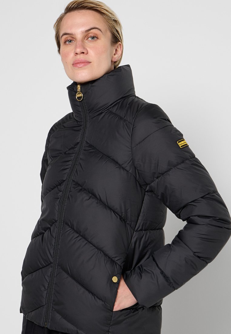 Schwarze gesteppte Pufferjacke mit hohem Kragen und goldenem Reißverschluss. Verfügt über diagonale Nähte und einen goldenen Akzent am linken Ärmel.