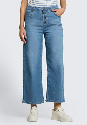 Femme portant un jean court large bleu clair avec quatre boutons à l'avant, une chemise rayée rentrée et des baskets blanches.