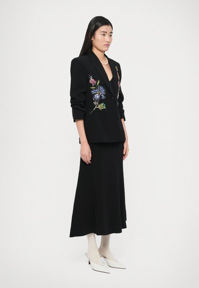 Fekete öltönykabát színes virágos hímzéssel, hosszú fekete szoknyával párosítva. A megjelenést fehér pántos magassarkú cipő és világos színű harisnya egészíti ki.