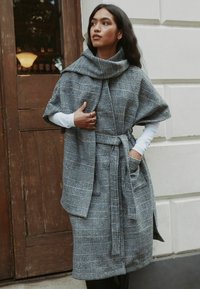 Hallidest ja mustadest ruudulistest kiududest moodustatud mantel kõrge krae, vööga talje ja suurte varrukatega, millel on taskud ja tekstuurse viimistlusega.