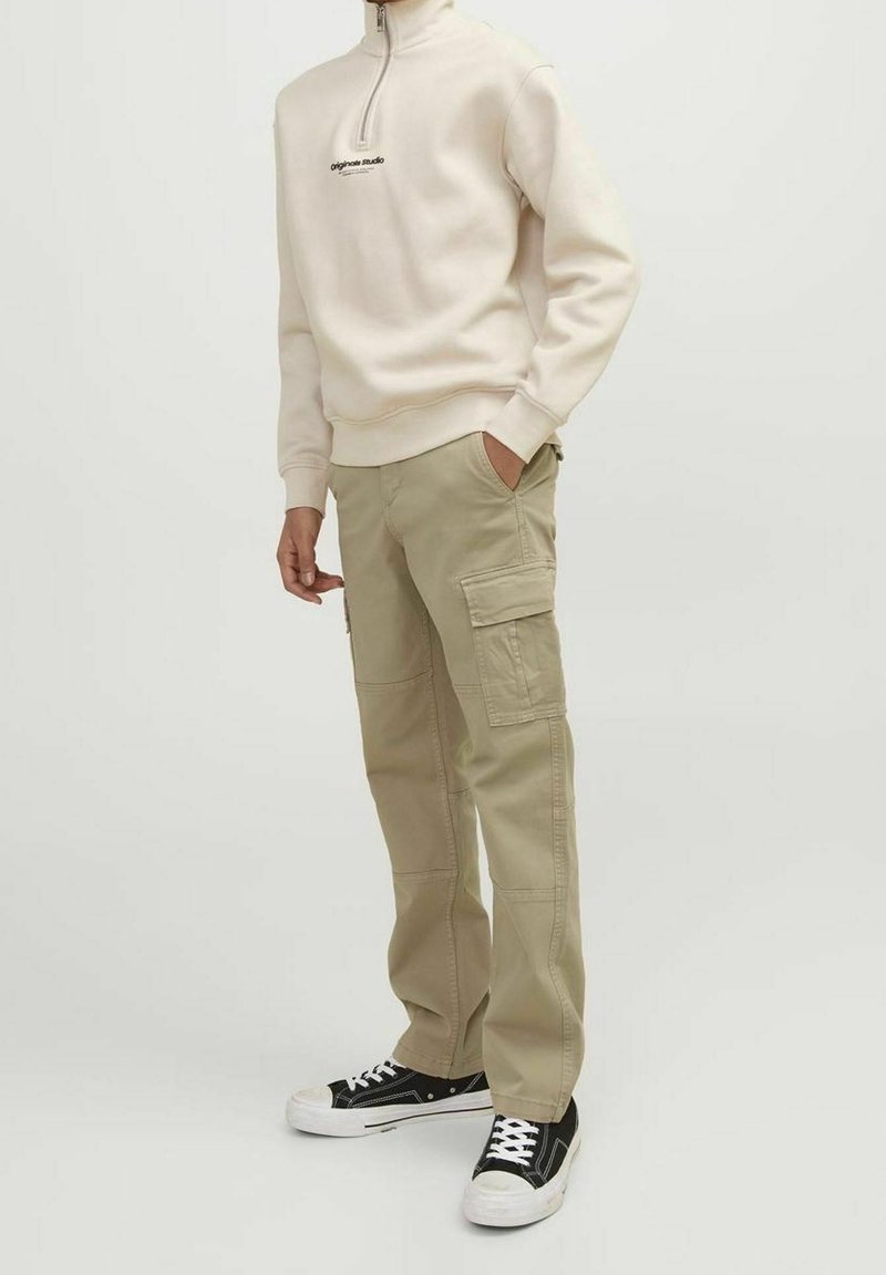 Homme portant un pantalon cargo beige, un sweatshirt crème à col zippé quart avec du texte, et des baskets noir et blanc, se tenant avec une main dans sa poche.