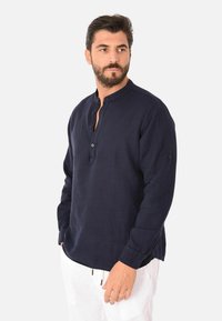 Camicia a maniche lunghe in lino blu navy con colletto rotondo, bottoni sulla parte anteriore, e polsini regolabili, indossata con pantaloni bianchi su uno sfondo neutro.