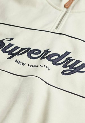 White sweatshirt with navy embroidered "Superdry" and "NEW YORK CITY" text. Features a zip closure and black piping details along the seams.