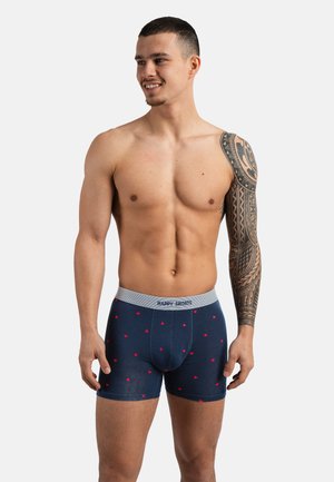 Smiling shirtless man with tribal tattoo sleeve wears navy boxer briefs with small red hearts and grey waistband labeled "HAPPY SHORTS."