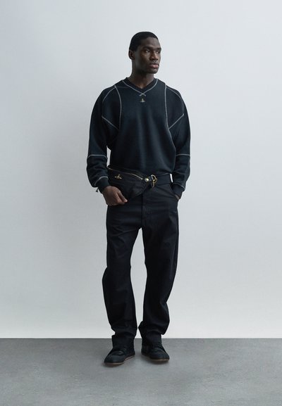 Άνδρας που φοράει μαύρο πουλόβερ με λευκή ραφή, μαύρο παντελόνι, μαύρα αθλητικά παπούτσια, κρατώντας μαύρο clutch τσάντα, στέκεται πάνω σε γκρι πάτωμα μπροστά σε λευκό τοίχο.