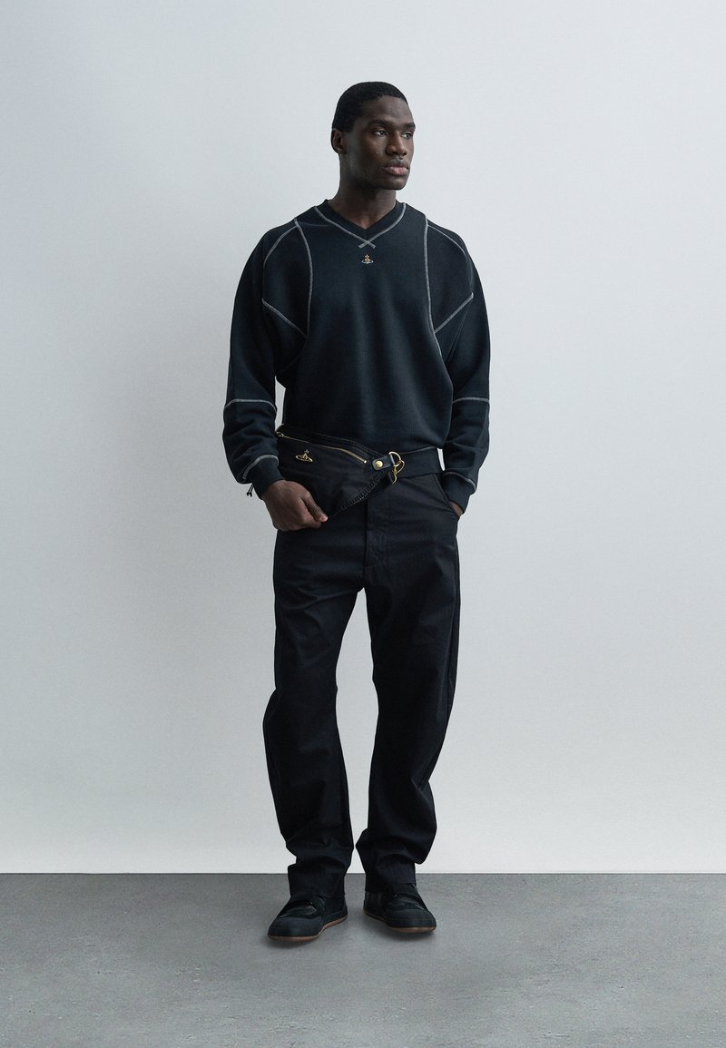 Uomo con maglione nero con cuciture bianche, pantaloni neri, scarpe da ginnastica nere, che tiene una pochette nera, in piedi su pavimento grigio contro parete bianca.