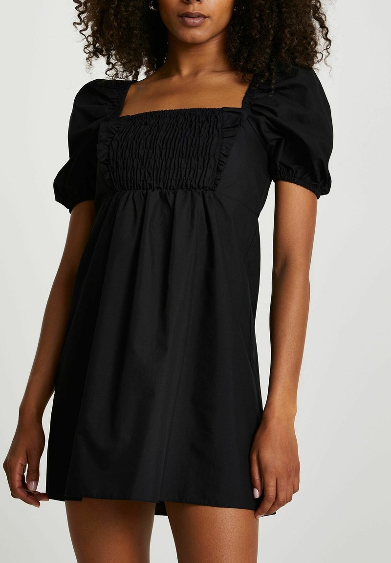 Femme portant une courte robe noire avec des manches bouffantes et un décolleté carré froncé, debout devant un fond uni.