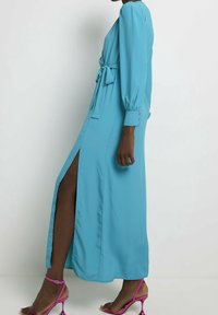 Dlouhý rukáv, tyrkysová maxi šaty se bočním rozparkem a páskem v pase. Materiál je měkký a rukávy jsou nařasené. V kombinaci s růžovými sandály na podpatku.
