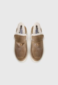 Pantofi slip-on din suede maro, cu căptușeală din blană sintetică albă și vârf rotund. Prezintă o curea cu brand pe partea superioară și o talpă crem.