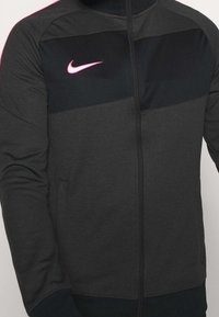 Dunkelgraue und schwarze Herrenjacke mit Reißverschluss, langen Ärmeln und einem pinken Nike-Swoosh-Logo auf der oberen Brust.