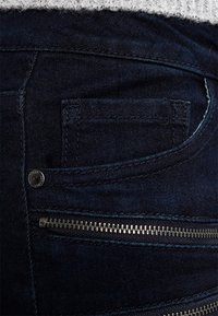 Los jeans de un índigo oscuro cuentan con un pequeño bolsillo trasero y un bolsillo con cremallera con herrajes metálicos. La tela tiene una textura suave y un brillo sutil.