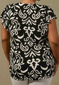 Svart topp med vitt blommönster, med volangärmar och avslappnad passform, visad bakifrån. Mjuk, texturerad tyg.