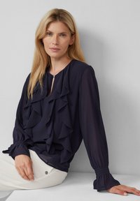 Blusa navy con dettagli arricciati sul davanti, tessuto leggero, maniche lunghe con polsini arricciati e scollatura a forma di chiave. Pantaloni bianchi visibili.