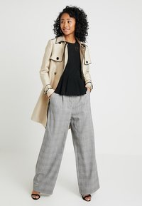 Beige trench coat over a black peplum top, paired with wide-leg plaid trousers in grey tones and black sandals. Curly hair framing the face.