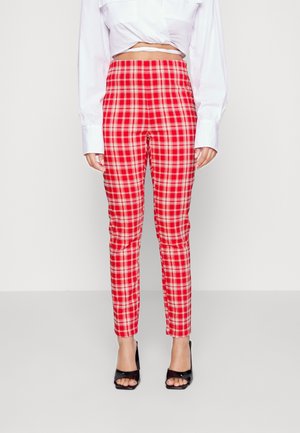 Kobieta ubrana w czerwono-białe wąskie spodnie w kratę z wysokim stanem, białą krótką bluzkę i czarne sandały na obcasie z odkrytymi palcami na jednolitym tle.