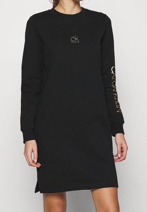 Mujer con un vestido negro de manga larga con el logo dorado de Calvin Klein en el pecho y la manga, posando frente a un fondo liso.