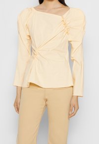 Kvinna iklädd en ljusgul blus med puffärmar och rynkade detaljer, ihop med beige byxor med hög midja.