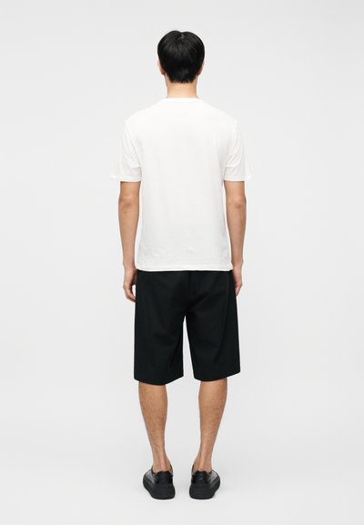 Άνδρας που στέκεται γυρισμένος με την πλάτη φορώντας λευκό απλό μπλουζάκι, μαύρα σορτς μέχρι το γόνατο και μαύρα παπούτσια χωρίς κορδόνια σε λευκό φόντο.