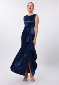 Robe satin marine sans manches, avec un devant drapé et une fente sur le côté. Tissu texturé avec une finition lisse. Sandales à talons transparentes.
