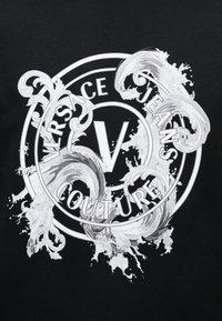 Λογότυπο Versace Jeans Couture με στυλιζαρισμένες λευκές βαρoκ σπείρες και πιτσιλιές χρώματος σε μαύρο φόντο.