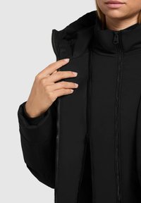 Osoba zapínající černou vyteplenou bundu s vysokým límcem, přičemž je zdůrazněn detail ruky a zipu proti jednolitému pozadí.